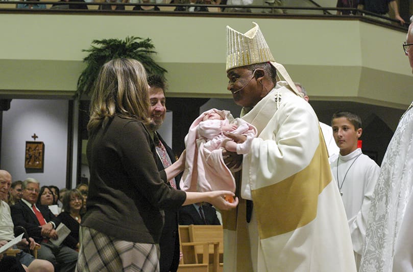 Archbishop Wilton D. Gregory holds one month old Kerri Wyglad as her parents Jenn and Kevin look on. They brought up the oil of baptism during the offertory procession. Photo By Michael Alexander