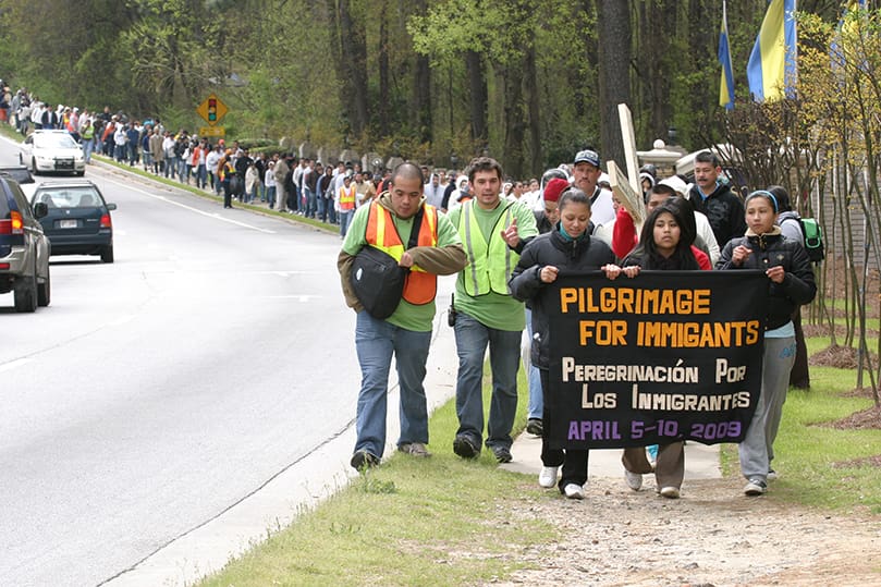 Mayra Garcia, left, and her sister Maria, right, carry the banner with Griselda Garcia (no relation) as the Interfaith Pilgrimage For Immigrants makes its way up Indian Trail-Lilburn Road in Gwinnett County. The top of the cross bears the name of the Roberto Martinez Medina, a 39-year-old Mexican citizen detainee at the Stewart Detention Center, Lumpkin, who died at St. Francis Hospital, Columbus, on March 11. While the results of an autopsy to determine the cause of death are still pending, questions surround the medical care for Medina and other detainee related deaths. Photo By Michael Alexander