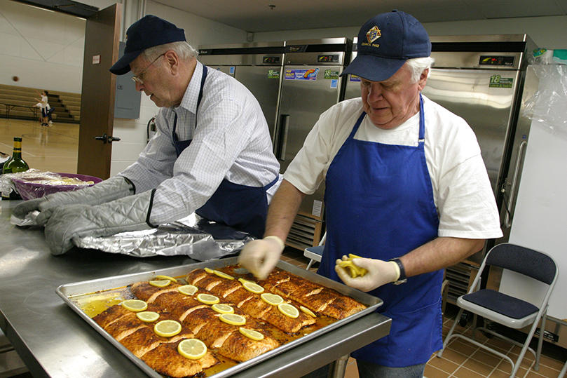 Andy Diaz, right, covers the cooked salmon with lemon wedges as Dave Webster wraps a tray of salmon, ready to go up to the serving area, with aluminum foil. Photo By Michael Alexander