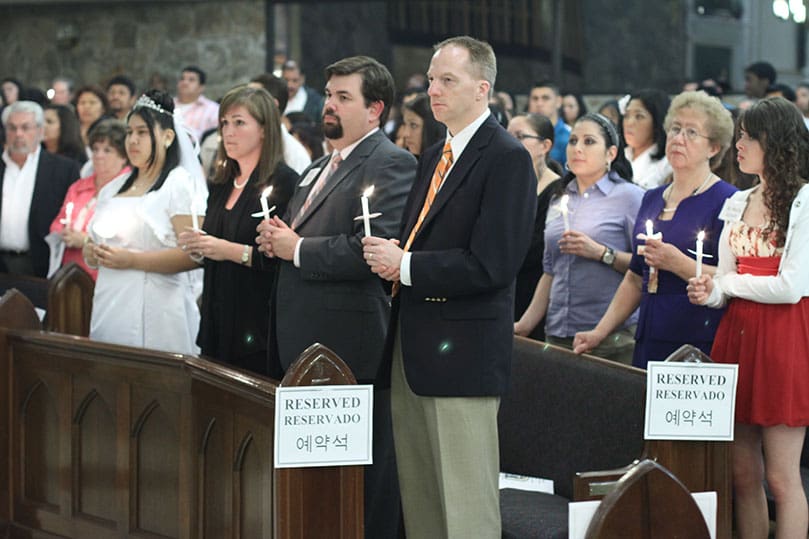 Standing in the church, the 26 candidates and their sponsors hold their miniature candles lit from the Easter Candle. Photo By Michael Alexander