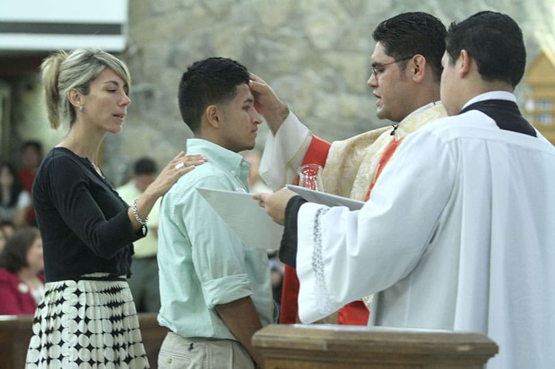 Eighteen-year-old Cesar Garcia, center, is confirmed by Father Refugio Oñate Melendez as his sponsor Cecilia del Toro, left, stands with him. Photo By Michael Alexander 