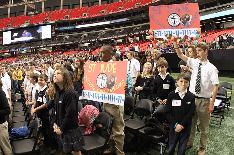 St. Jude the Apostle School eighth-graders Chandler Parks, center, and Jack Gillett hold signs as the students watch the paper chains unfurl and rise to heights above the stage on both sides. Through various acts of compassion and kindness displayed by Rachel’s Challenge participants, they estimate that 10 miles of chains were collected and brought to The Georgia Dome. Photo By Michael Alexander