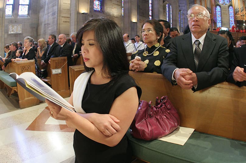 Teresa Thuy Nguyen, kneeling foreground, the older sister of ordination candidate Tri Nguyen, joins the congregation in prayer during the Litany of the Saints. In the pew immediately behind her are their parents Mary Thao, left, and Trong. Photo By Michael Alexander