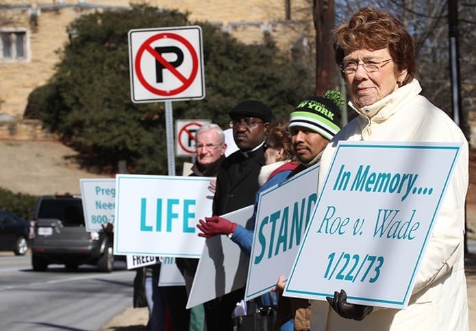 Lenore Schrowang, foreground right, stands along Peachtree Road, just above West Wesley Road, with her pastor Father Henry Atem and other fellow parishioners from St. George Church, Newnan.  Photo By Michael Alexander