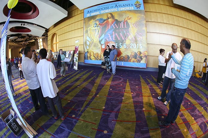 As one couple demonstrates, the 15 by 15 foot, 2015 Eucharistic Congress banner hanging in the north ballroom lobby provides a popular backdrop for camera phone portraits as attendees walk by it during the 20th annual Eucharistic Congress at the Georgia International Convention Center, College Park. Photo By Michael Alexander