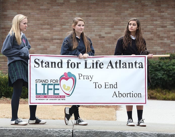 St. Pius X High School students (l-r) Grace Berg, Mallory Wood and Nikki Castro hold a sign as they stand on Peachtree Road in front of the Cathedral of Christ the King, Atlanta. Photo By Michael Alexander