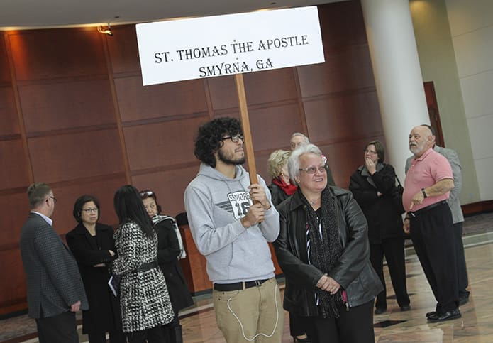 Marco Gonzalez, 18, of St. Thomas the Apostle Church, Smyrna, holds a sign in the Cobb Galleria Centre atrium bearing the parish name, so other candidates like him, catechumens and sponsors can be directed to the St. Thomas the Apostle seating area. Standing with Gonzalez is RCIA program volunteer Lauren Russell. Photo by Michael Alexander