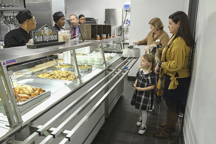 As principal Lisa Cordell, background right, passes out utensils to students, Sienna Smith is the first student to go through the food line, accompanied by her kindergarten teacher Karen Cosse. The new cafeteria was up and running for students and teachers on Nov. 29. Photo By Michael Alexander
