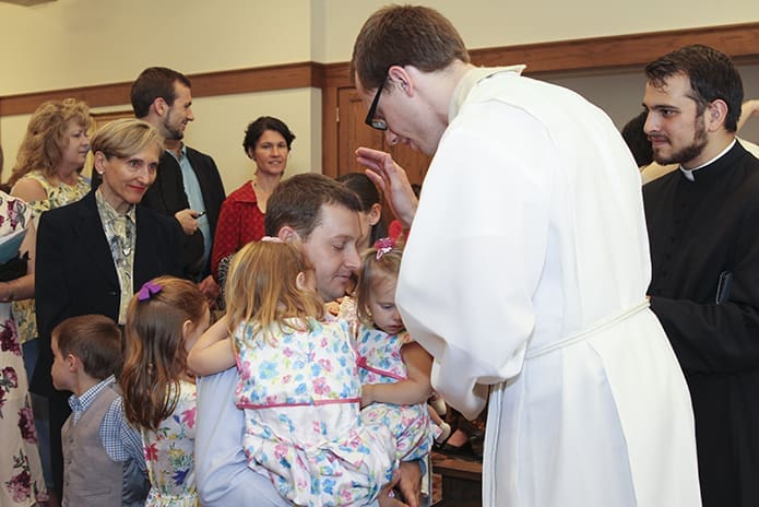 Father Michael Bremer extends a blessing to Jeff Graebner as he holds two of his children, three-year-old Mary, left, and two-year-old Christina. Photo By Michael Alexander
