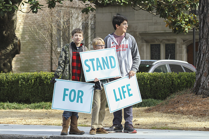 (L-r) Will Zeliff, 12, Ben Gallamore, 13, and Christian Gonzales, 13, stand along Peachtree Road beside the Cathedral of Christ the King, Atlanta, with their signs facing the traffic going north. Zeliff, Gallamore and Gonzales attend Our Lady of the Mountains Church, Jasper, Christ Redeemer Church, Dawsonville, and Good Shepherd Church, Cumming, respectively. Photo By Michael Alexander