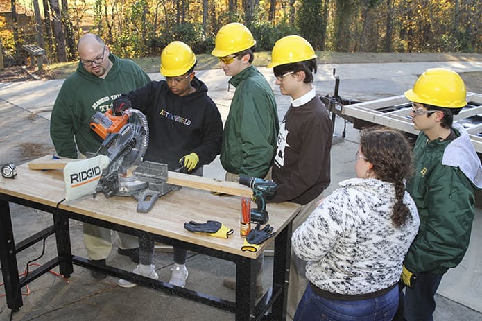 (Counterclockwise, from right) Senior Rosie Nemec, sophomore Brendan McDonnell, and juniors David James and Luca Cella look on as junior Marcus Solomon saws a piece of wood. Dr. Brent Hollers, far left, project manager and business and technology education teacher, also observes. It was the second build day, Nov. 28, of the school's tiny home project. Photo By Michael Alexander