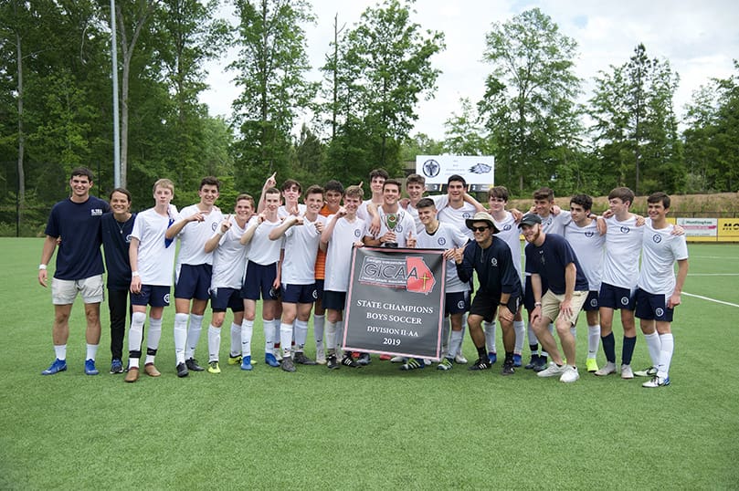 St. John Bosco Academy's soccer coaches (in dark T-shirts, l-r) Alex Brenner, head coach, and assistant coaches Claire Sullivan, Alex Sullivana and Michael Houlihan celebrate their state championship with the players during a group photo. Photo Courtesy of St. John Bosco Academy