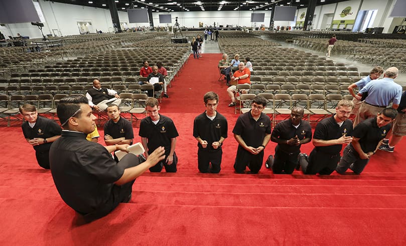 Father Rey Pineda, foreground left, gives some instructions to a group of seminarians, kneeling, as they rehearse their parts for various aspects of the 2018 Eucharistic Congress. In the background, permanent deacons and laity in roles of responsibility sit and stand in the vast hall at the Georgia International Convention Center. Once the doors are open to the public, that space is transformed into the English and Spanish tracks. Photo By Michael Alexander