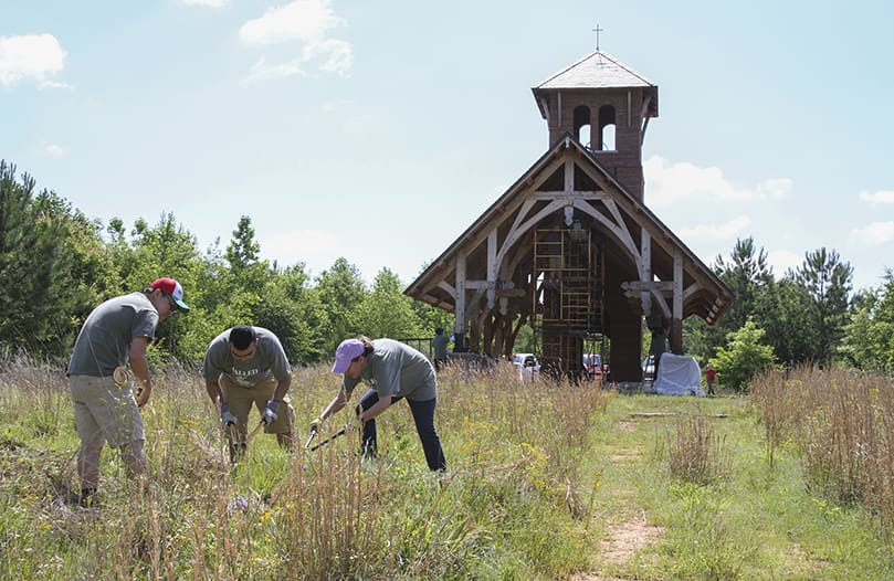(Left to right) Seminarian Joseph Nguyen, Jonathan Patino, a young man in discernment, and Sally Scardasis, executive assistant to the vocations director, trim the brush around burial plots as other seminarians work on staining the Martin Gatins Chapel in the background. It took place during a May 16 seminarian workday on the natural burial grounds of the Monastery of the Holy Spirit’s Honey Creek Woodlands, Conyers. Photo By Michael Alexander