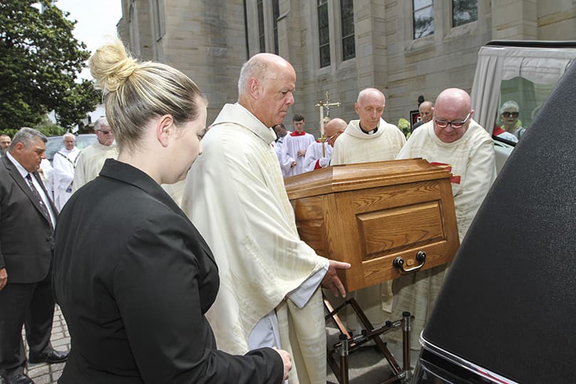 Elana Ray, a funeral director for H.M. Patterson & Son - Spring Hill Chapel, Atlanta, looks on as three of the six pall bearers (clockwise, from left), Msgr. Peter Rau, pastor of St. Peter Chanel Church, senior priest Father John Adamski and Msgr. Francis McNamee, rector of the Cathedral of Christ the King, Atlanta, place Father Richard Morrow's casket in the funeral hearse. Photo By Michael Alexander