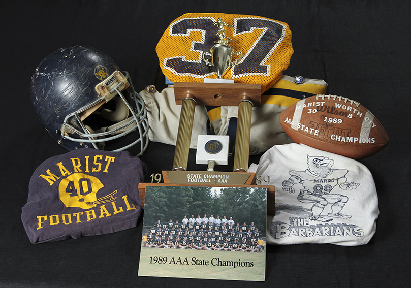 A program, with a photo of the 1989 Marist School football team on its cover, rests at the base of the Class AAA championship trophy, which is on top of some “battle grey” football pants. Other items from that year include championship rings, team jerseys, a game ball with the championship game’s final score, the helmet of a former player and a T-shirt bearing the nickname of the ’89 team for its aggressive style of play. Photo By Michael Alexander