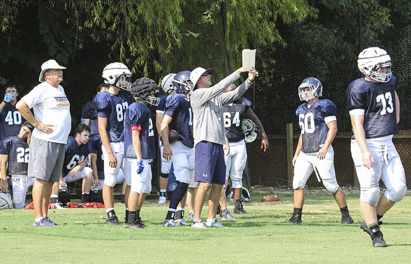 Offensive line coach Daniel Perez, center, points out a play to some players during an August afternoon practice. Linebacker coach Gary Miller, left, looks on from behind. Perez and Miller are the only current coaches, along with head coach Alan Chadwick, that were around in 1989 when the Marist football team went undefeated (15-0) and won its first state championship. Perez also played quarterback for Marist in 1982 and 1983. Photo By Michael Alexander