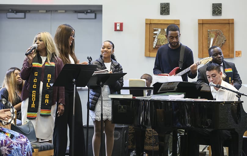 (Standing, l-r) Rochelle Calhoun sings the musical prelude, “We Worship You,” with Lyke House choir members Nebechi Okpala, a Georgia State University junior, Lila Gilliam, a Spelman College freshman, and Morehouse College seniors James Floyd and Gabriel Savage, seated at the piano. Jarron Okin, far right, playing the tambourine, is a member of the Ghanaian Catholic Community Choir. Calhoun, in town for the anniversary Mass, was a cantor at Father Urey Mark’s former St. Louis, Missouri parish, St. Nicholas.