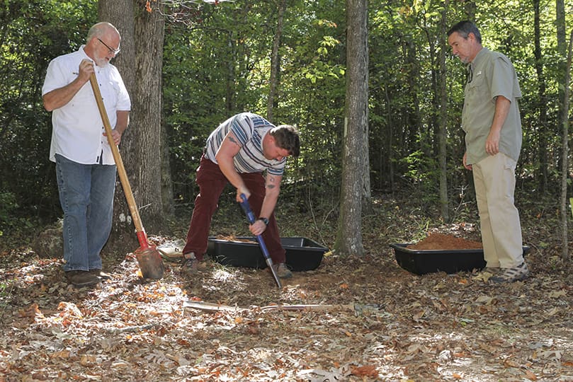 (L-r) Michael Wojcik, steward at Honey Creek Woodlands, Conyers, and Jeff Brennan take turns digging a hole for an upcoming double cremation service. Looking on from the right is retired manager and burial marker engraver, Joe Whittaker. Photo By Michael Alexander