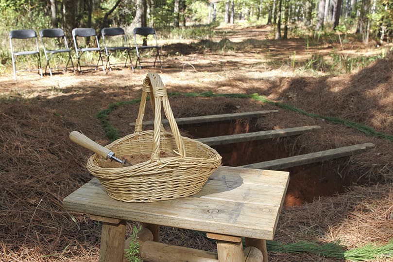 The scene is nearly set for an afternoon burial in the Pine Forest section of Honey Creek Woodlands, Conyers. In order to preserve their freshness, flowers are one of the last things placed at the burial site. On the table, in the foreground, is a basket of ceremonial dirt and a garden trowel. It’s there if family members would like to toss some dirt over the deceased person’s burial plot. Photo By Michael Alexander