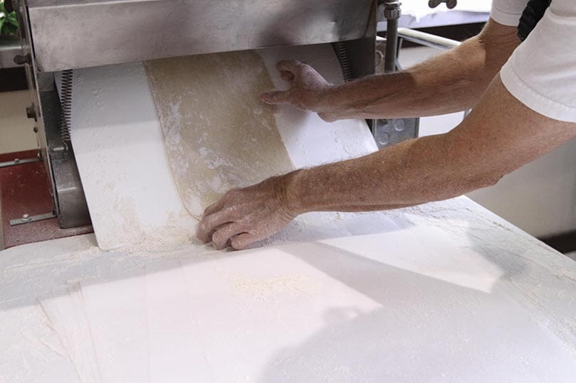 After going through the machine, the dough comes out underneath. At that point, several completed ones are carried into the kitchen for the men working around the table. Photo By Michael Alexander
