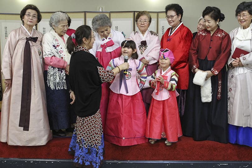 Connie Pyo, holding the microphone, served as mistress of ceremonies during a Lunar New Year celebration that followed the 10 a.m. Mass at St. Andrew Kim Church, Duluth, Jan. 26. Six-year-old Alexis Turner shares some words as a fellow youngster and senior females of the parish take the stage wearing the traditional Korean dress known as a hanbok. Photo By Michael Alexander