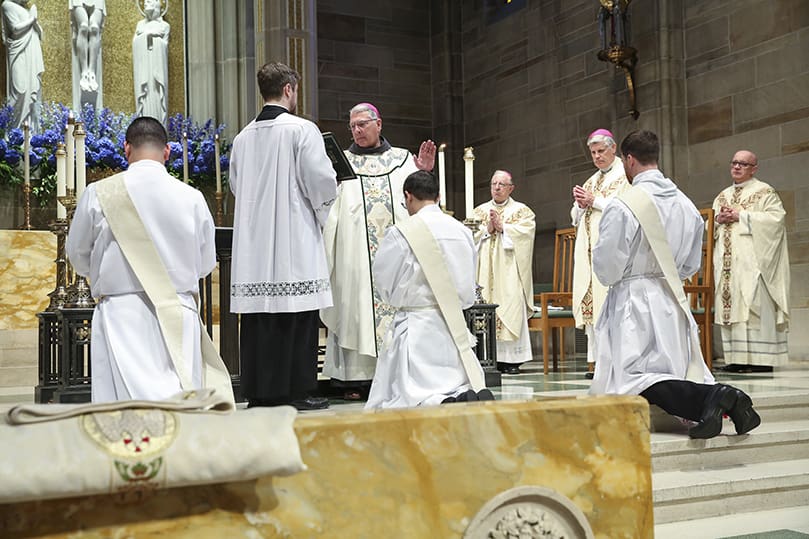 Archbishop Gregory J. Hartmayer, OFM Conv., standing, second from the left, conducts the prayer of consecration. At its conclusion, the three transitional deacons officially become priests. Photo By Michael Alexander