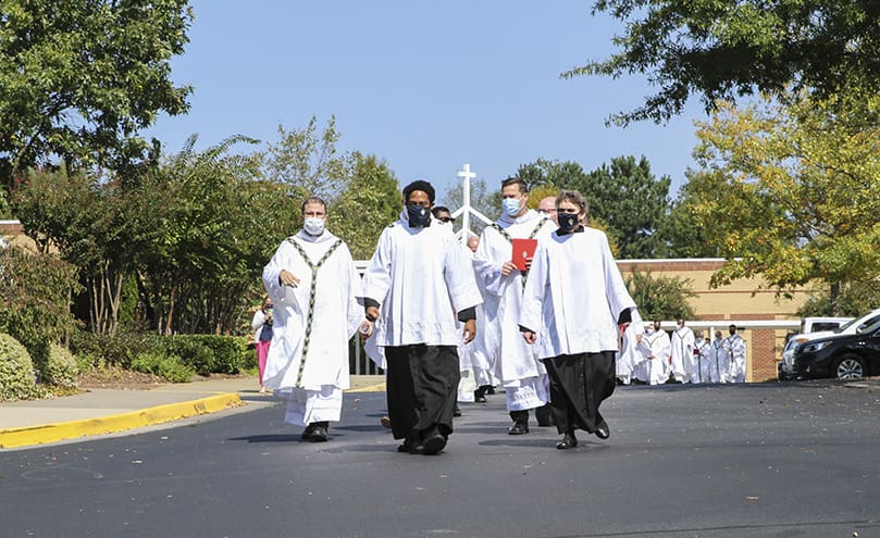Seminarian and master of ceremony Avery Daniel, left, and the Office for Divine Worship’s Patricia DeJarnett, right, lead the procession of priests to St. Peter Chanel Church for the pallium Mass. Photo By Michael Alexander