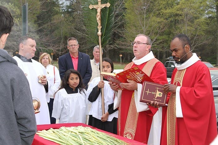 Deacon Larry Welsh of St. Matthew Church, Winder, second from right, reads the Gospel of Luke 19:28-40 on Palm Sunday as they stand before the congregation gathered in front of the church. Standing with him is the pastor Father Gilbert Exumé. Photo By Michael Alexander