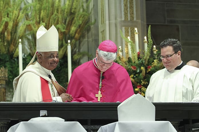 With the two of the three insignias of his office, the bishop’s ring and miter, in the foreground, and a smiling Archbishop Wilton D. Gregory, left, standing at his side, Bishop-designate Bernard E. (Ned) Shlesinger III signs the oath of fidelity and the formula for the Profession of Faith. Looking on is Father Joshua Allen, Georgia Tech Catholic Center chaplain, and one of the masters of ceremonies for the vespers service. Photo By Michael Alexander