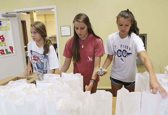(L-r) Kaylee Buckley. Mary Hays, and Jessica Tardy, rising sophomores at St. Pius X High School, Atlanta, fill lunch bags with pretzels and cookies at their table station before they are moved on for other items like juice and sandwiches. Photo By Michael Alexander