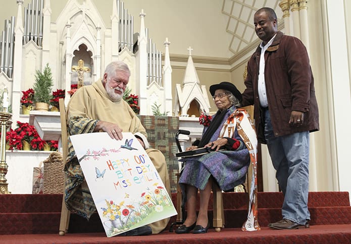 Msgr. Henry Gracz, left, pastor of the Shrine of the Immaculate Conception, Atlanta, presents Emily Milner with a poster size birthday card. Standing behind her is Melvin Denham of Orlando, Fla., her youngest grandson. Photo By Michael Alexander