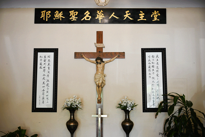 A crucifix hangs on the wall to the entrance of Holy Name of Jesus Mission with flowers on each side. The Chinese symbols above spell  “Holy Name of Jesus Chinese Catholic Church”.  And on the sides, “Because God did not send his Son into the world to judge the world but to save the world. God so loved the world that he gave his only begotten Son so that whoever believes in him will not perish but have eternal life.” Photo by Julianna Leopold