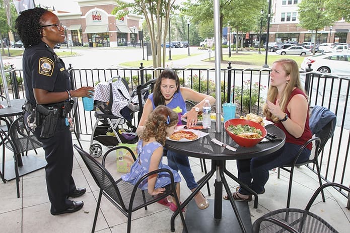 Brookhaven Police Department pedestrian safety officer Maria Jones, standing left, returns to the outdoor patio table at Newk’s Eatery, where she shared lunch with (r-l) Our Lady of the Assumption Church youth minister, Angela O’Donoghue, Erin Rogan and her 3-year-old daughter, Audrey. Photo By Michael Alexander