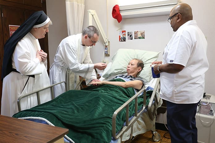 Hawthorne Dominican Sister Damien, left, and licensed practical nurse (LPN) Leroy John, right, stand by as Father Paul Burke gives holy Communion to cancer patient Khon Tran. Each morning following Mass, Father Paul and Sister Damien visit all the patients, who either receive Communion, a blessing or both. Photo By Michael Alexander