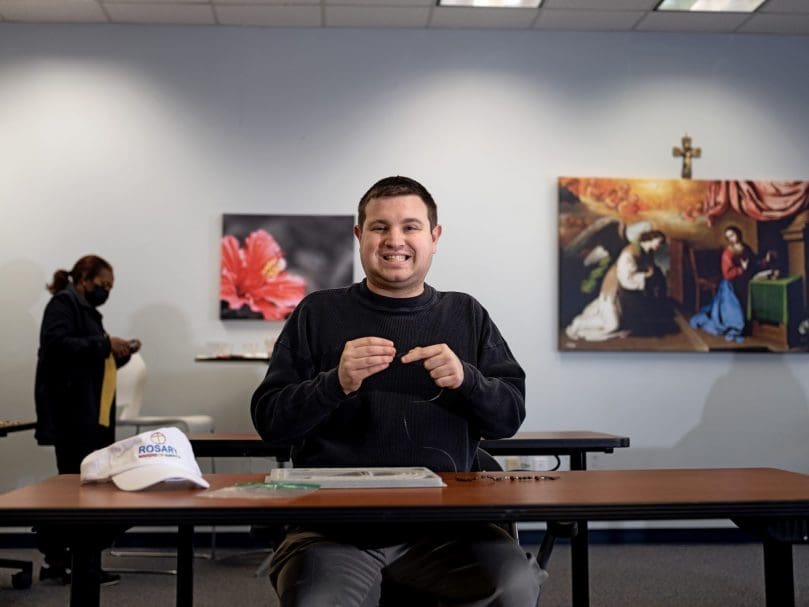 Michael Feliu pauses work to share a smile while making a rosary at the Rosary Makers of America office. Photo by Johnathon Kelso