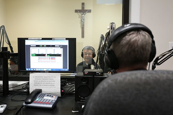From the other side of the glass, Father Luis Guillermo CÃ³rdoba, background, administrator of Our Lady of the Americas Mission, converses with Bishop Luis R. Zarama, foreground, during programming of the Spanish Catholic radio program, âNuestra Fe.â  Photo By Michael Alexander