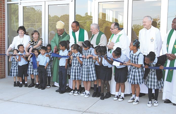 Archbishop Wilton D. Gregory cuts the ceremonial ribbon in front of the new Enrichment Center at St. John the Evangelist School, Hapeville as (back row, l-r). Diane Starkovich Ph.D., superintendent of schools, St. John the Evangelist School principal Karen Vogtner, Father Michael Onyekuru, current pastor of St. John the Evangelist Church, former pastor Msgr. Edward J. Thein, currently rector of the Basilica of the Sacred Heart of Jesus, Atlanta, Franciscan Father John Koziol, pastor of St. Philip Benizi Church, Jonesboro, former pastor Father Richard Morrow and Scalabrinian Father Jacques Fabre, administrator of San Felipe de Jesus Mission, Forest Park, look on with children from the school. Photo By Michael Alexander