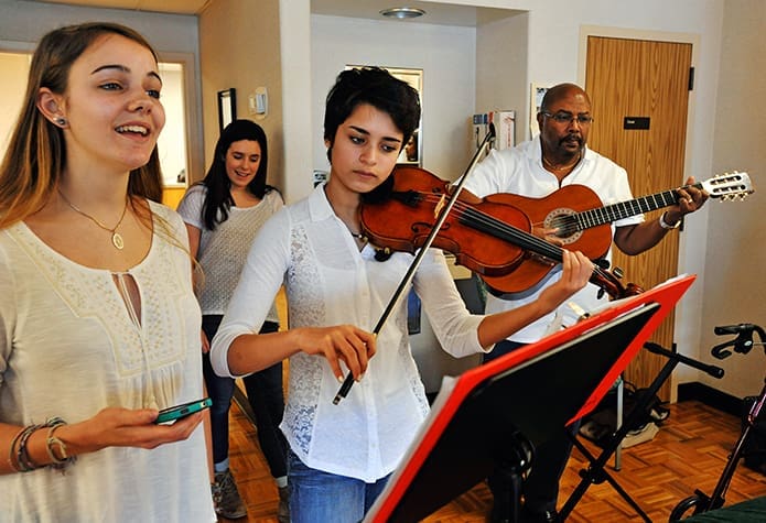 Caroline Tanzy, 17, from St. Pius X High School and Michaela Pocock, 17, on violin, from Roswell High School are joined by staff member Leroy John on guitar, in a musical performance for the patients at the cancer home. Singing along in the background is 16-year-old Maggie Weir, also from St. Pius X High School. Photo By Lee Depkin
