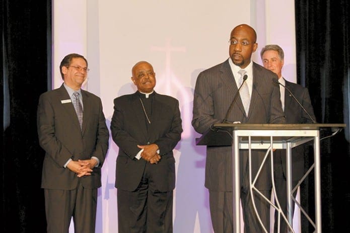 Rev. Dr. Raphael G. Warnock, senior pastor, Ebenezer Baptist Church, addresses the Catholic Charities Gala audience. Rev. Warnock was awarded the 2013 Archbishop Donoghue Award. Looking on in the background are (l-r) Catholic Charities Atlanta executive director Miguel San Juan, Archbishop Wilton D. Gregory and Catholic Charities Atlanta chairman of the board, Scott Garrett.