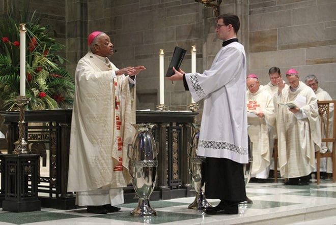 Archbishop Wilton D. Gregory, left, leads a prayer during the consecration of the chrism as seminarian Michael Metz holds the Roman Pontifical.