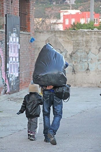A Syrian refugee and his son walk to the bus that will take them to a border camp. Formerly middle class, this father carries all of the possessions gathered over a lifetime in a black plastic bag after being forced from his home by war. Photo By Mikaele Sansone/Catholic Relief Services
