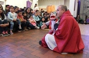 Father Fabio Alvarez Posada, sitting right, holds Father Fa, one of two puppets that lend a helping hand in bringing the message of Jesus Christ to the children in his parish. Photo By Michael Alexander